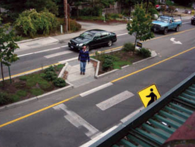 A person waits to cross a narrow road on a well-designed pedestrian-friendly median